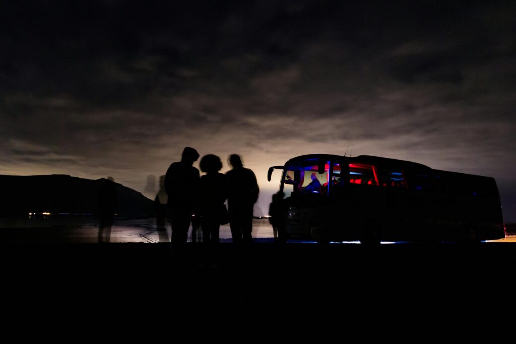 Silhouetted figures and a bus under Iceland's night sky create a mysterious coastal scene.